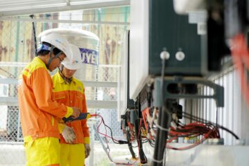 Two technicians with helmets working on electrical equipment outdoors, ensuring safety. (Photo by Trinh Trần on Pexels)