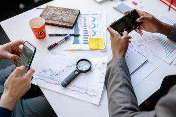 Two professionals analyzing data and graphs with smartphones on a white table. (Photo by Yan Krukau on Pexels)