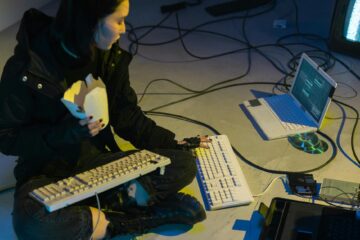 A young woman in a dark room surrounded by computers and cables, eating and typing on keyboards. (Photo by cottonbro studio on Pexels)