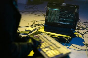 A person typing on a keyboard connected to a laptop showing code, surrounded by wires. (Photo by cottonbro studio on Pexels)