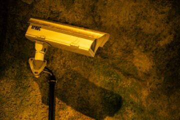 Close-up of a security camera mounted on a textured wall at night. (Photo by Patrick on Pexels)