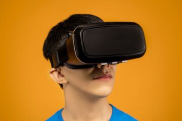 Young man immersed in virtual reality using VR headset against a vibrant orange background. (Photo by Sound On on Pexels)