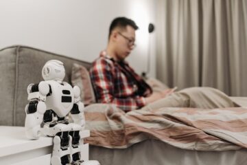 A modern bedroom featuring a robot on a bedside table and a man sitting on a bed. (Photo by Pavel Danilyuk on Pexels)